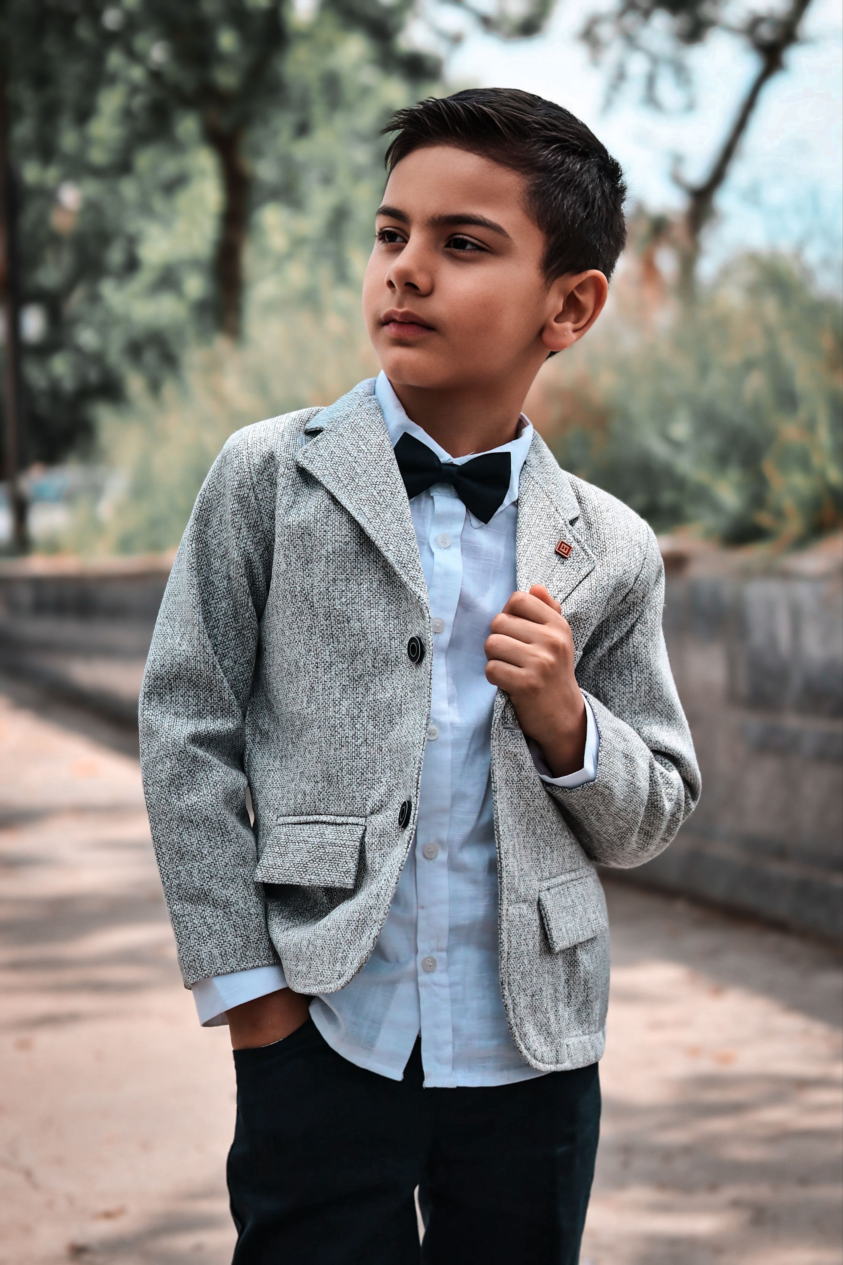 Um garoto vestido com um elegante terno cinza e gravata borboleta, em pé, confiante, ao ar livre.