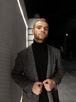 Young stylish man in a grey coat posing outdoors at night, exuding confidence and fashion flair.
