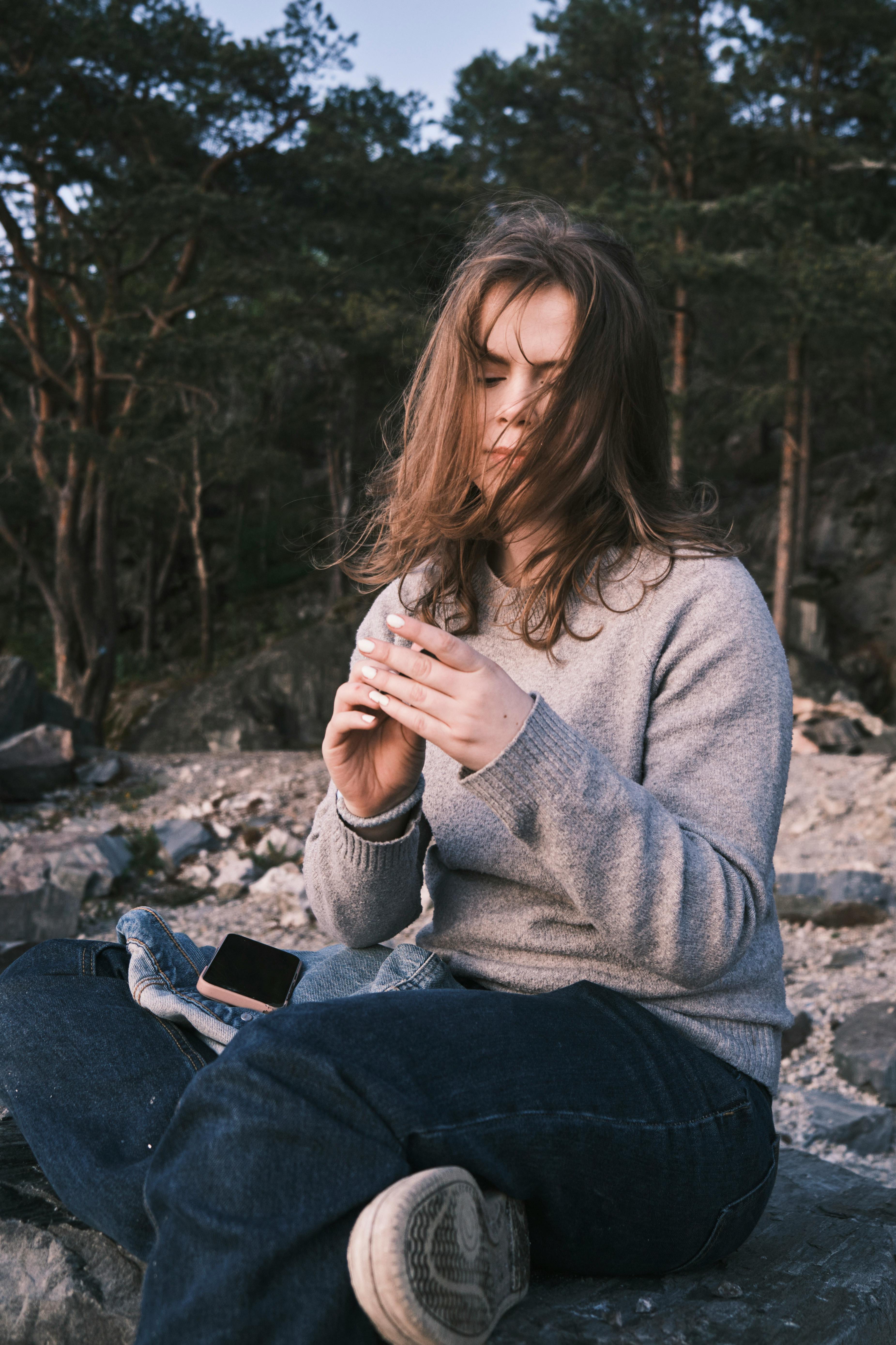 Young woman with phone sitting on a rocky ground in a forest setting.