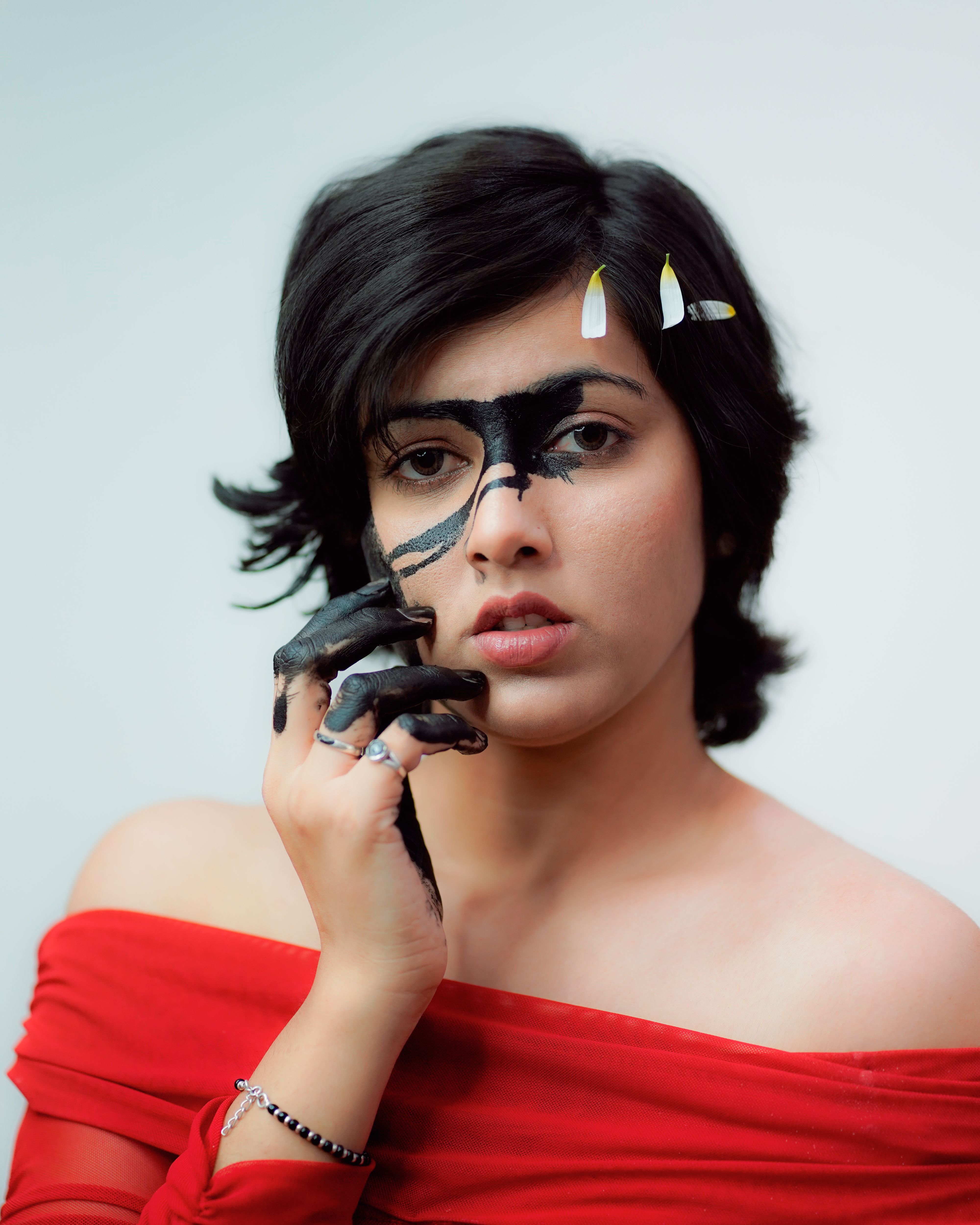 Artistic portrait of a woman with black face paint, unique makeup, and a red off-shoulder top, creating a striking visual.