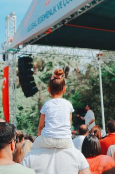 A child sitting on an adult's shoulders at an outdoor festival with a stage in the background.