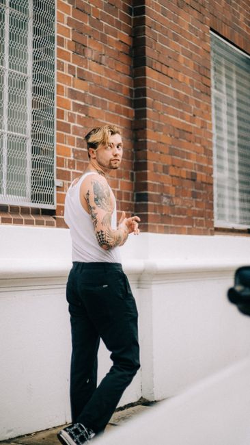 A fashionable tattooed man casually posing against a brick wall outdoors, showcasing style and confidence.