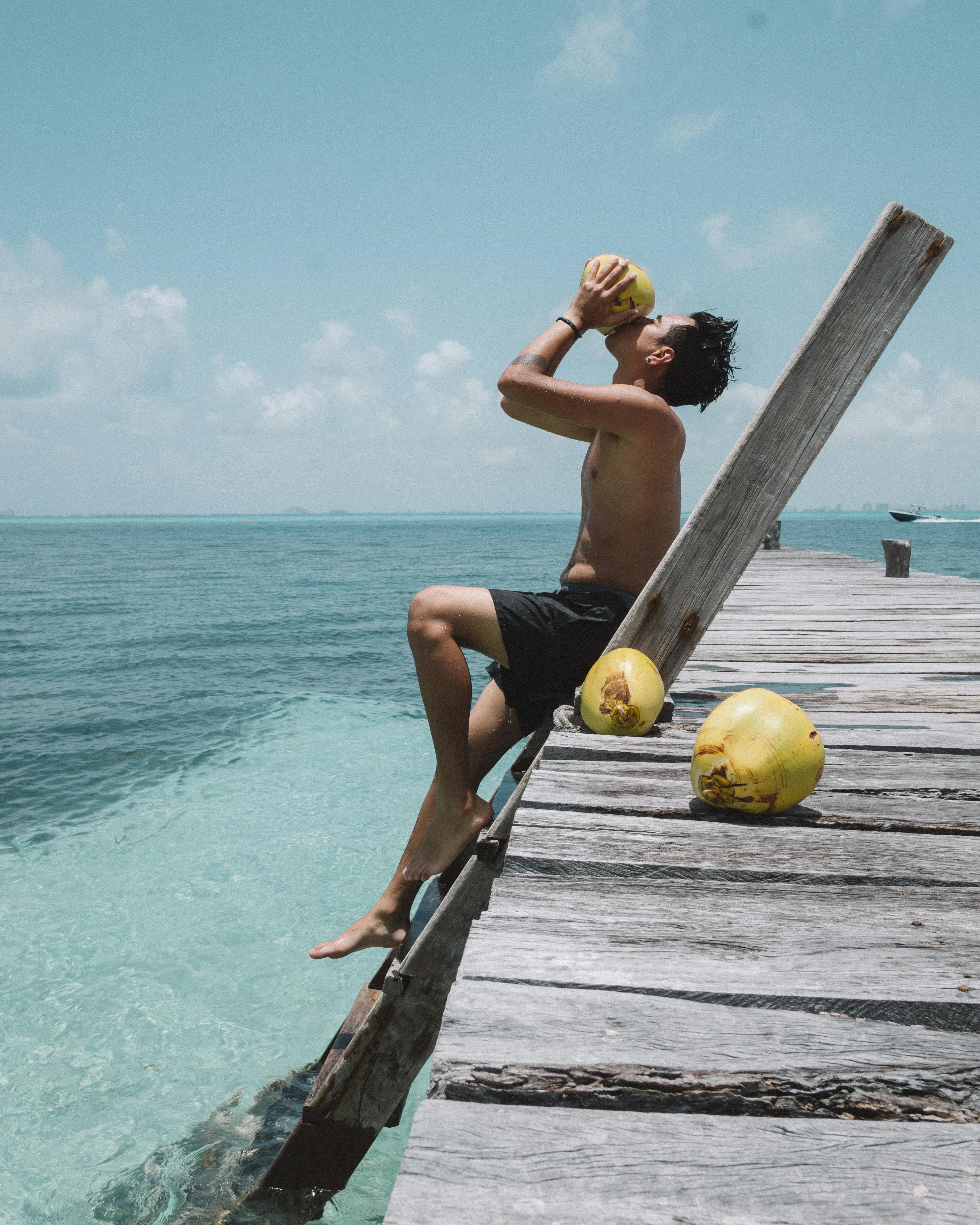 Shirtless man drinking coconut on wooden pier at Isla Mujeres, Mexico.