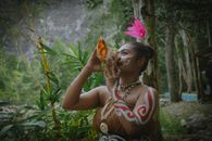 A woman in tribal attire plays a conch shell in a forest setting, capturing cultural heritage.
