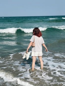 Woman enjoying ocean waves barefoot on a sunny beach, holding shoes.
