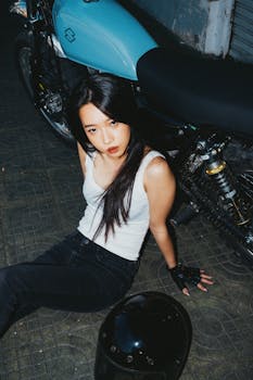 Stylish young woman sitting by a blue motorcycle with a helmet at night.