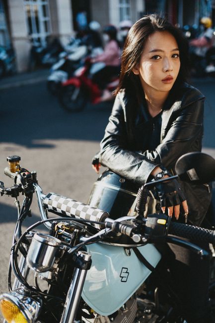 Young woman in leather jacket posing confidently on a motorcycle in a city street.