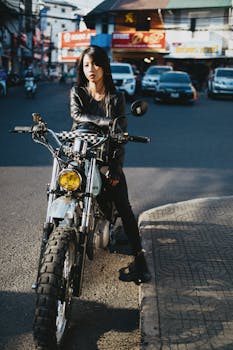 A confident woman in black clothing sits on a motorcycle in a bustling urban street.