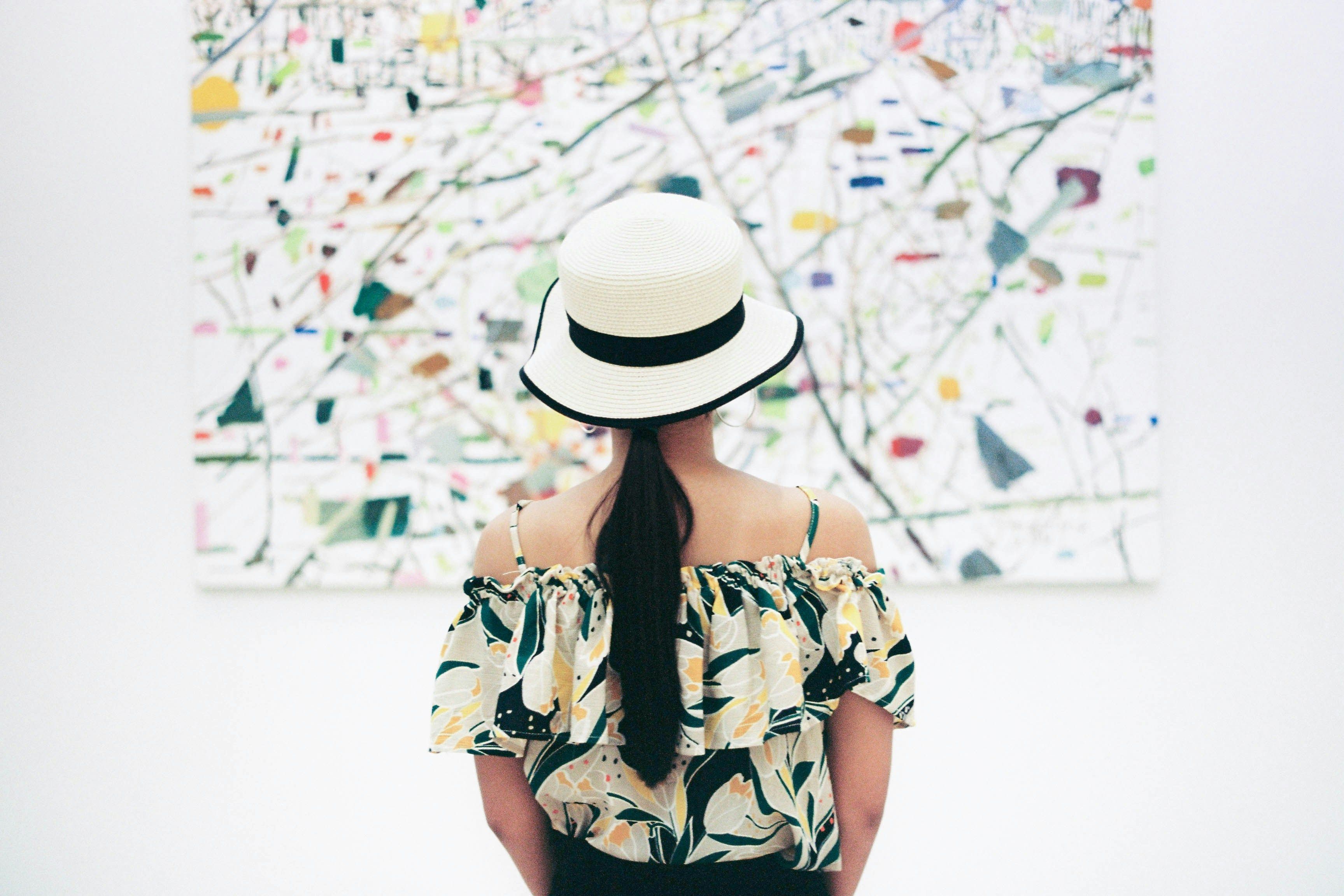An Asian woman with a hat observes abstract art in a museum gallery, highlighting cultural exploration.