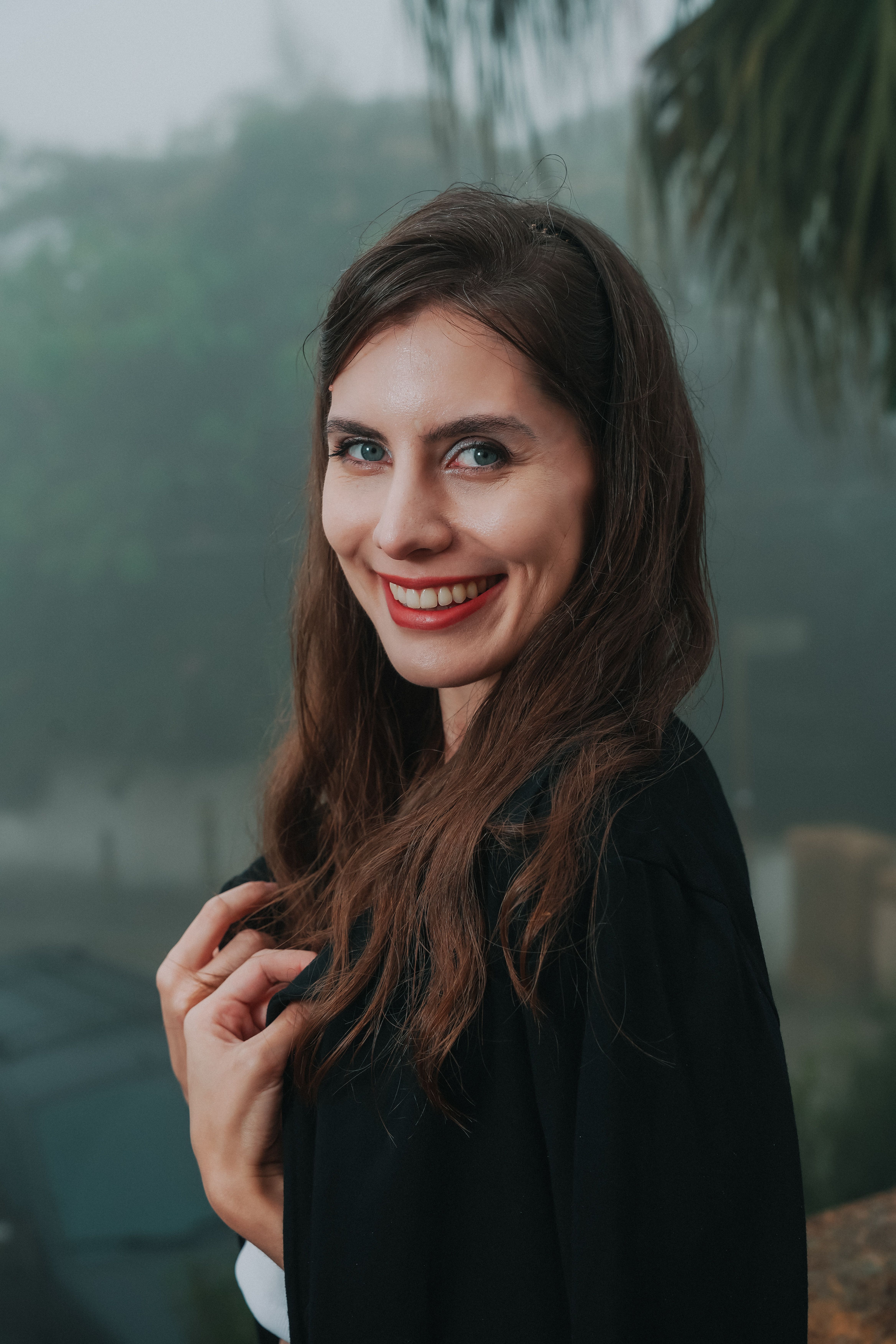 A woman with long brown hair smiles warmly, outdoors on a misty day.