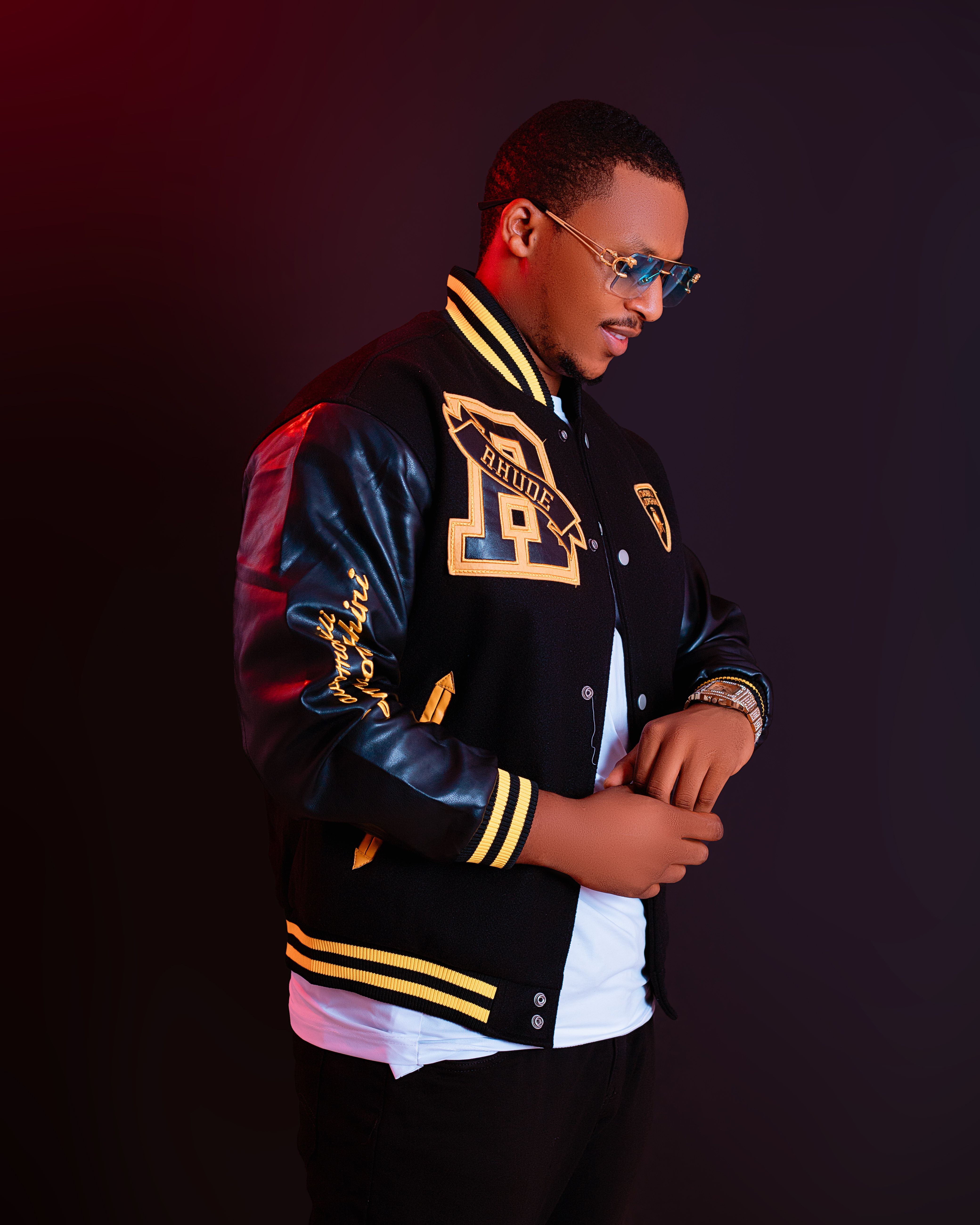 Young man wearing a trendy varsity jacket and sunglasses against a dark backdrop.