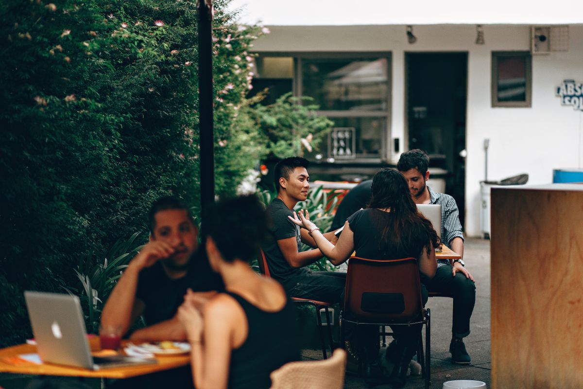 People talking around a table with laptops at an outdoor cafe — photo by Helena Lopes on Pexels