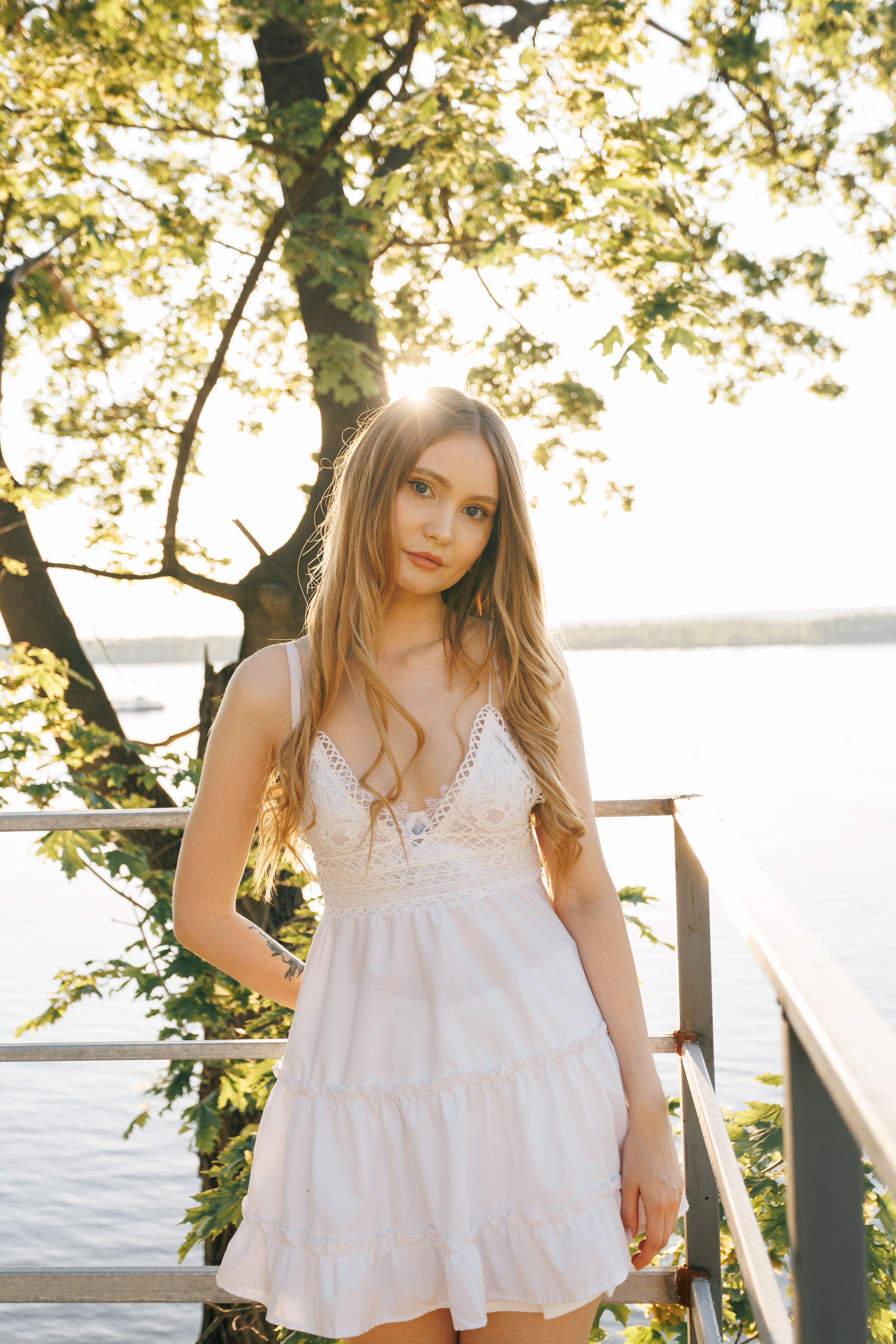 Young woman in a white dress standing by a lake in summer, sunlight filtering through trees.