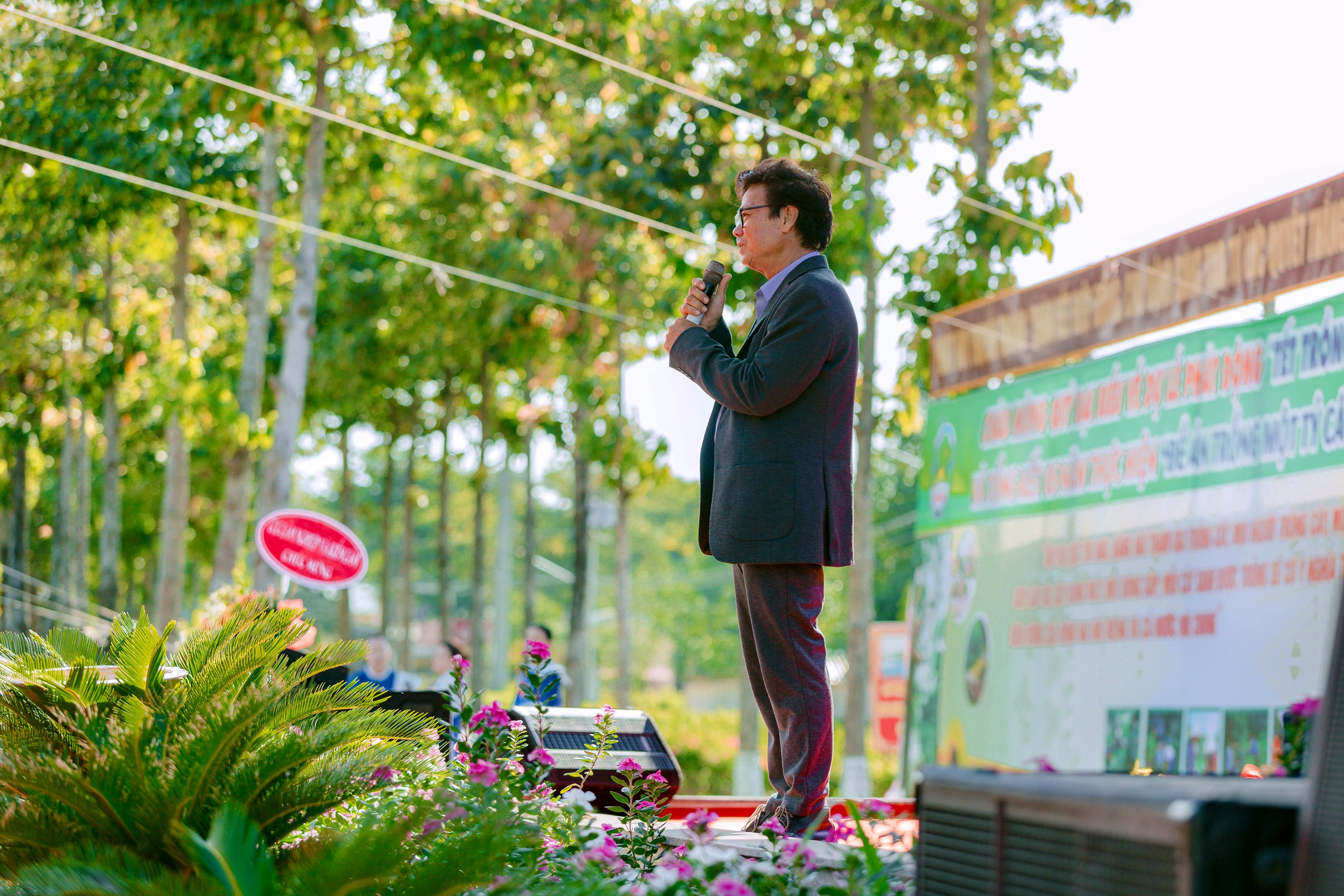 Man giving a speech at an outdoor event surrounded by trees and plants.