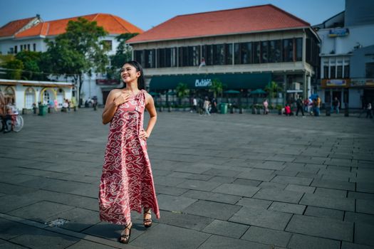 Asian woman wearing a stylish batik dress poses confidently in Jakarta's vibrant city square.