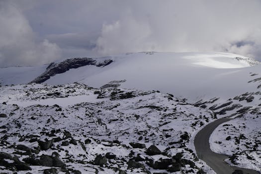 A tranquil winter landscape featuring a winding road through snowy mountains, under a cloudy sky.