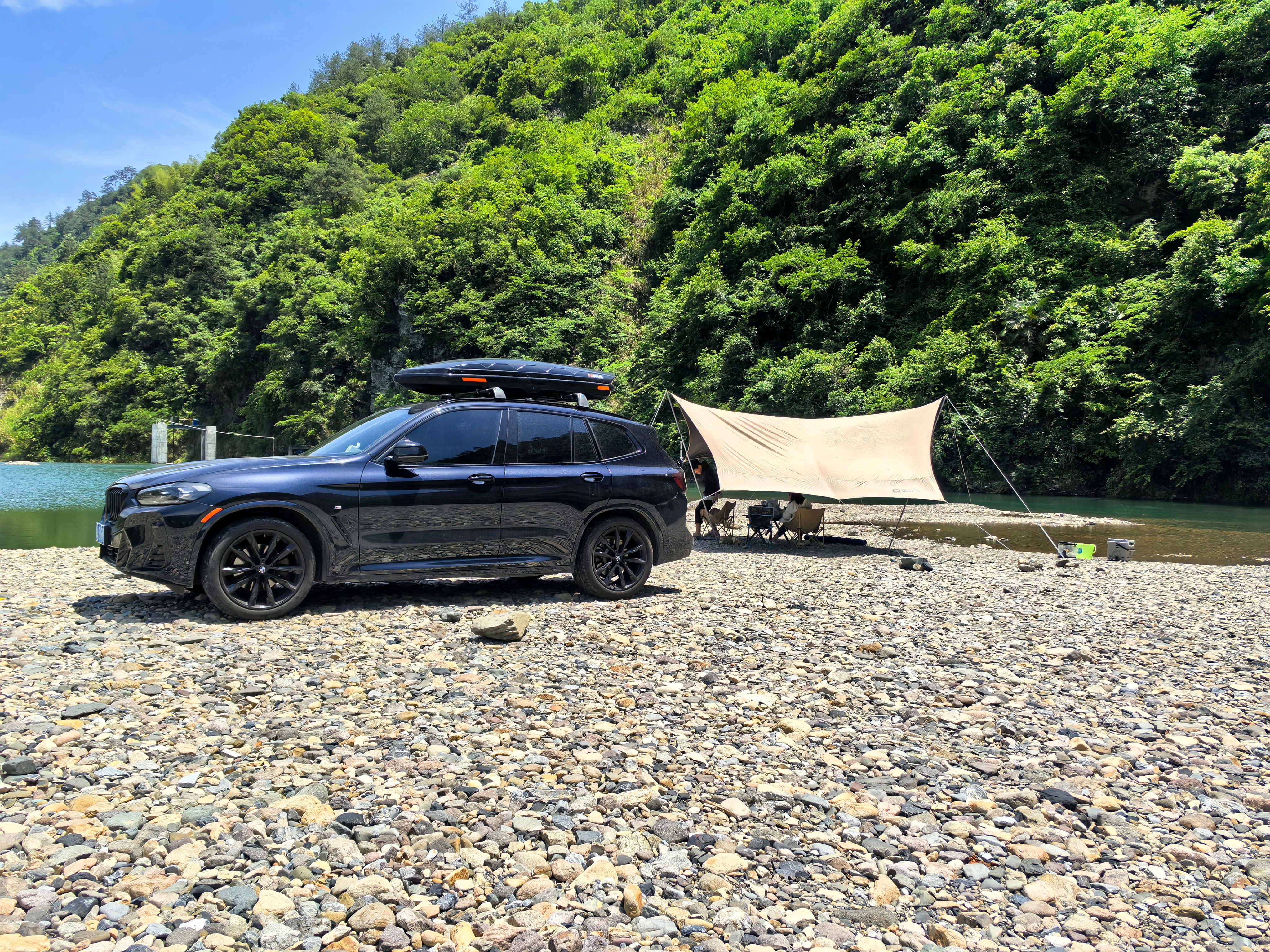 A black SUV parked beside a scenic river with a camping setup under a tent.