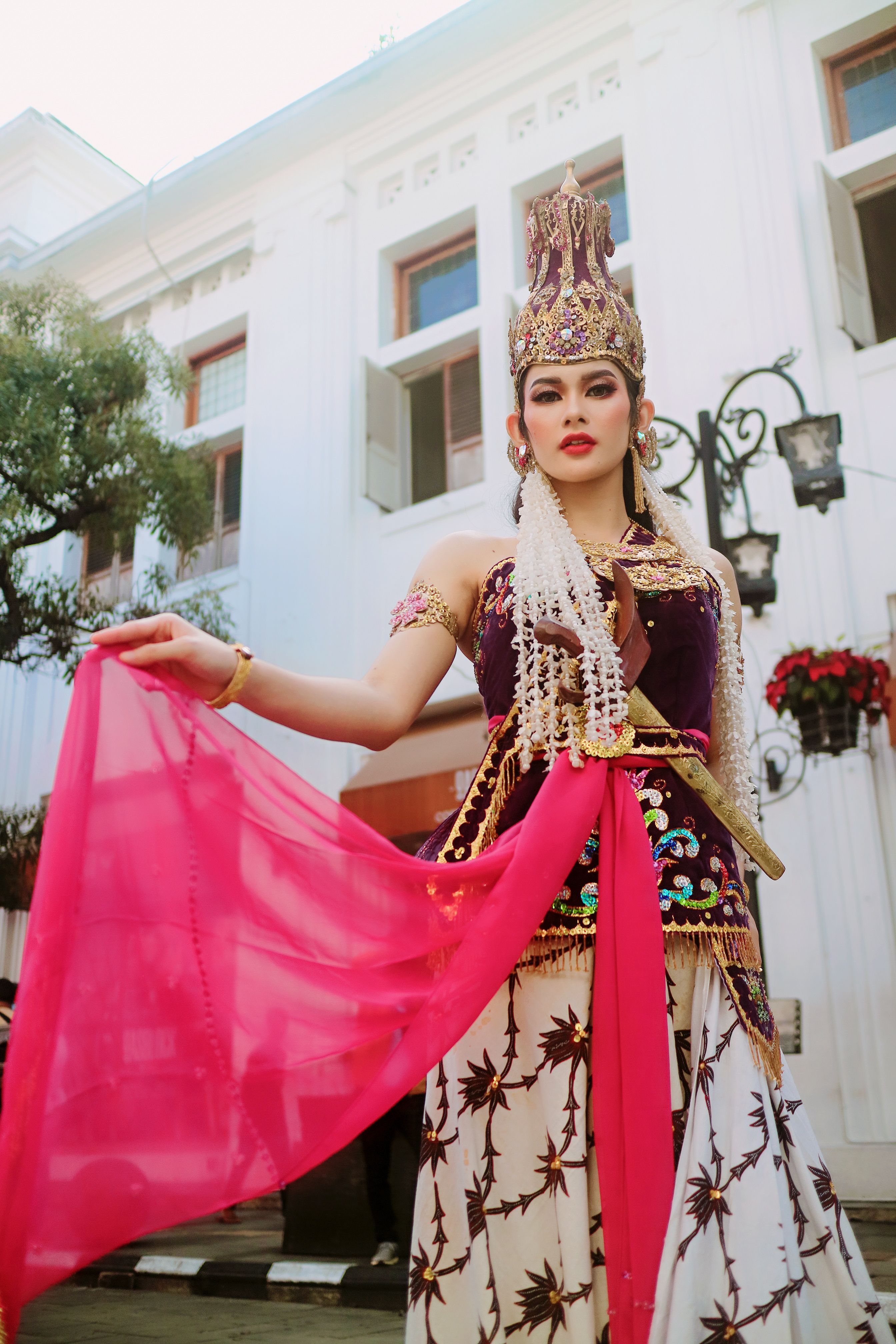 A woman in traditional Indonesian attire poses outside a historic building, alluring elegance and cultural pride.