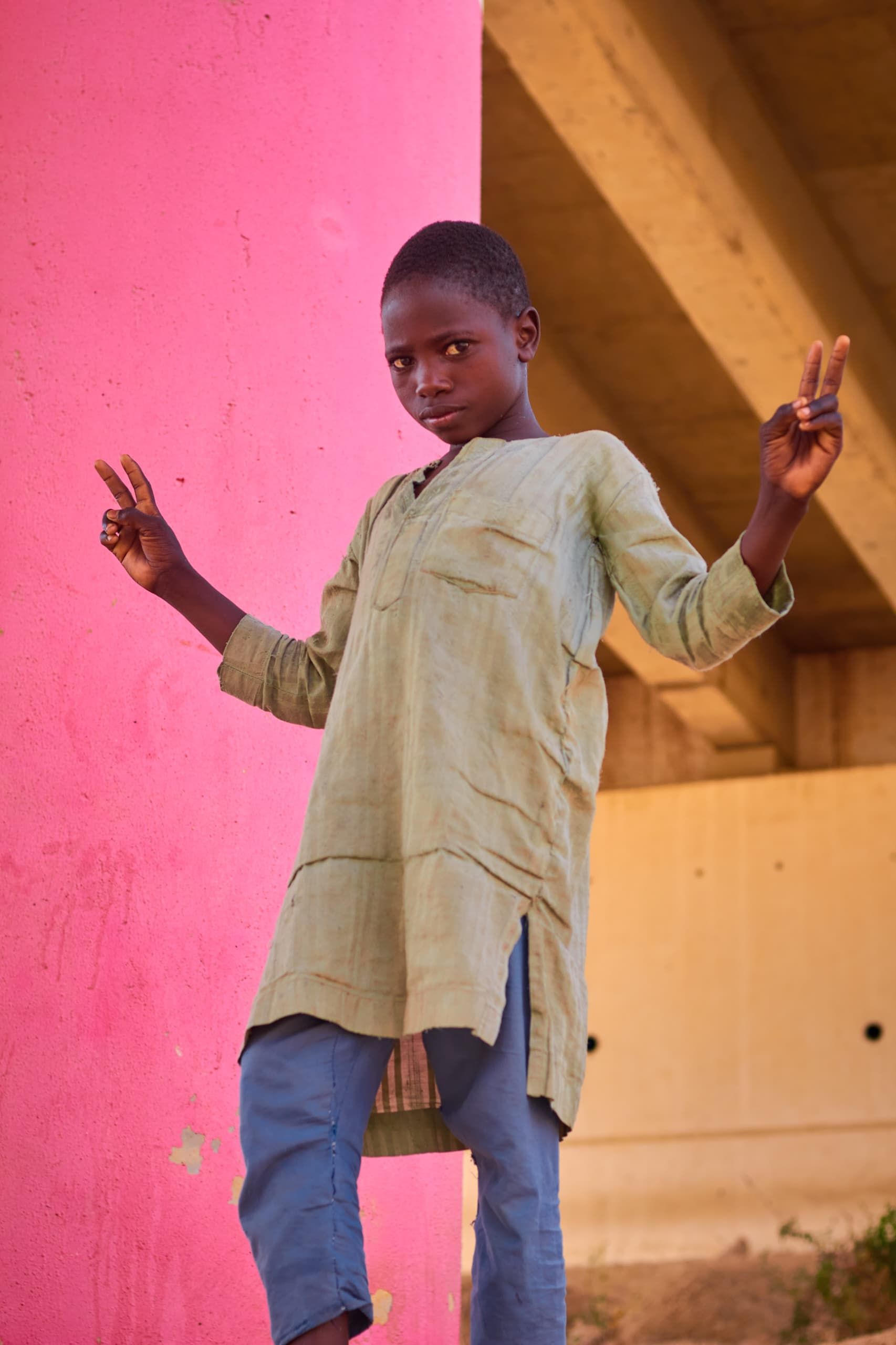 A young boy shows peace signs in front of a vibrant pink wall, expressing joy and playfulness.