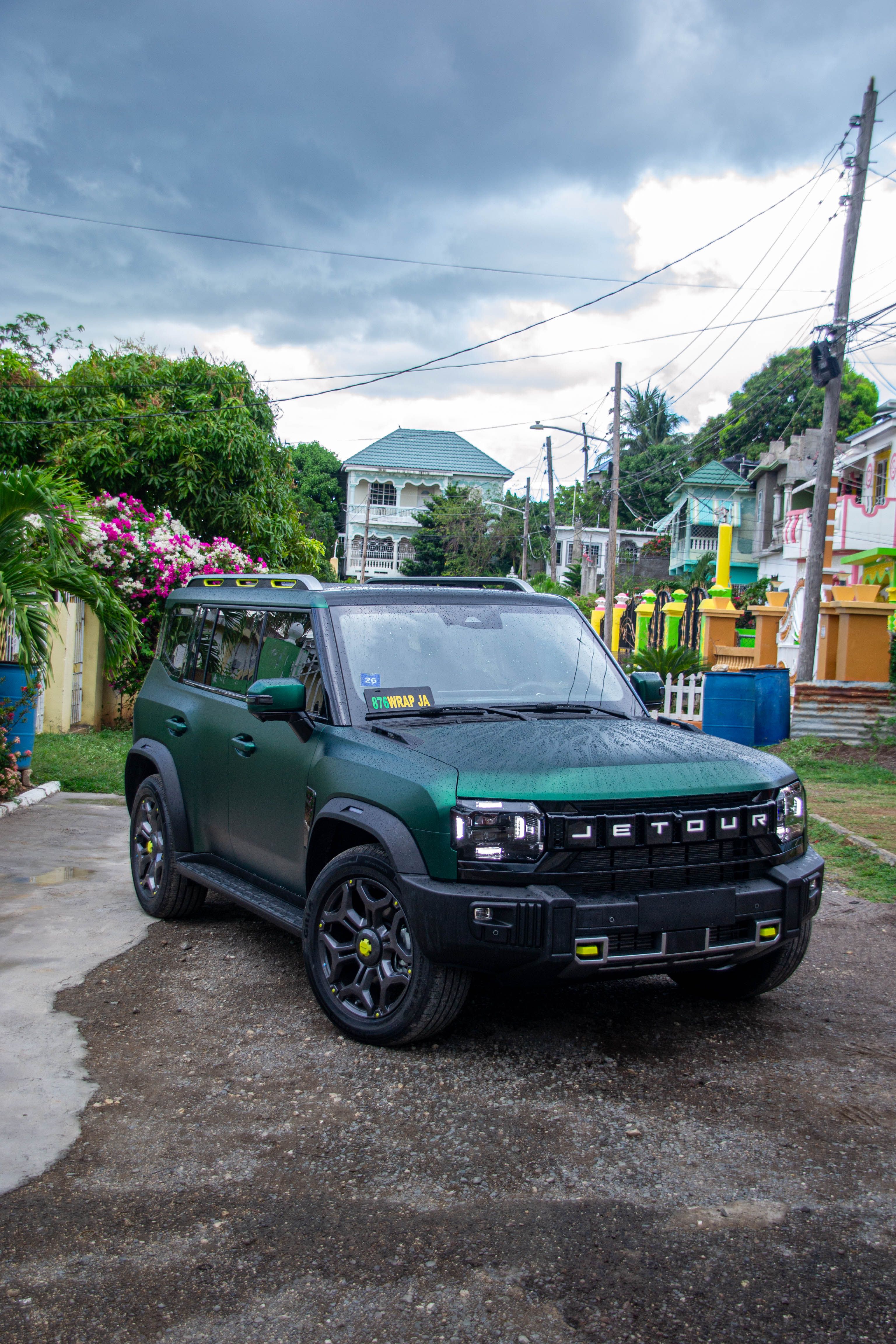 A green Jetour SUV is parked on a residential street with cloudy skies above and colorful houses in the background.