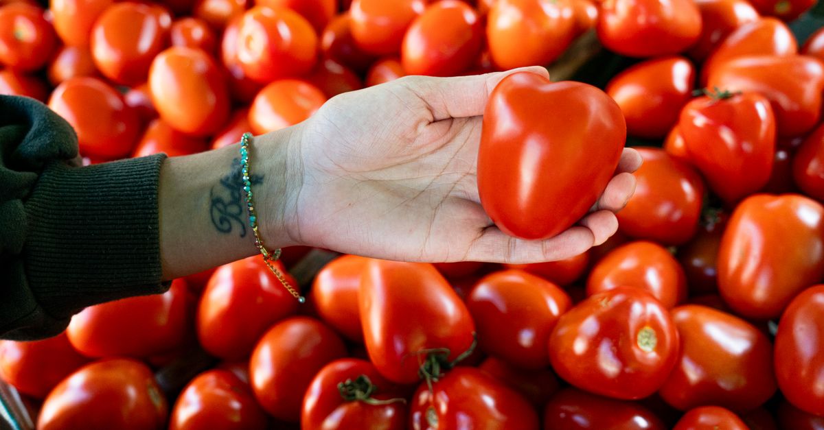 Hand holding a unique heart-shaped tomato among a pile of fresh produce.