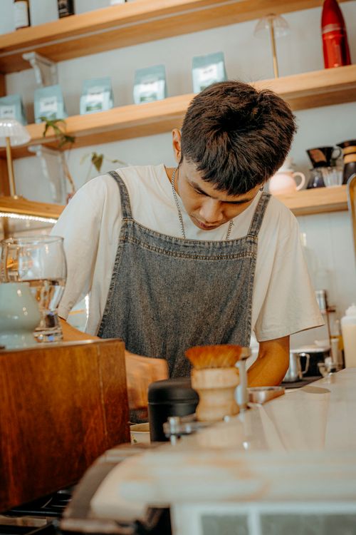 Free Barista in denim apron preparing coffee in a cozy cafe with shelves in the background. Stock Photo