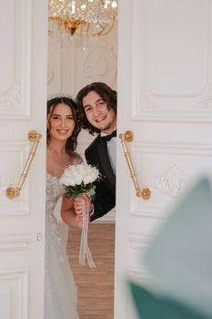Smiling bride and groom peeking through ornate doors. Elegant setting.