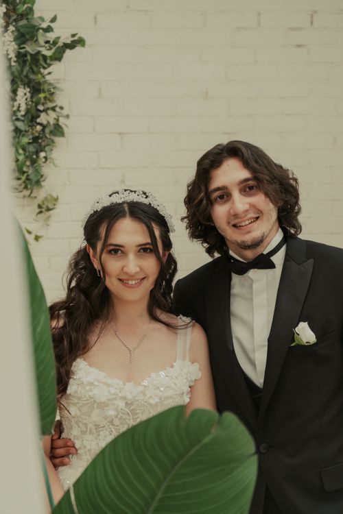Free A joyful couple in bridal attire posing indoors with a leafy backdrop, celebrating their wedding day. Stock Photo