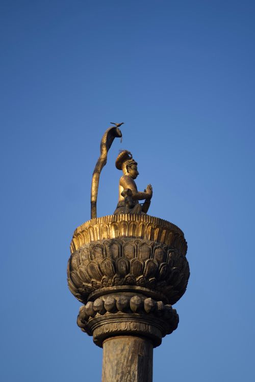 Free A golden statue depicting a prayerful figure atop a carved pillar with a clear blue sky background in Nepal. Stock Photo