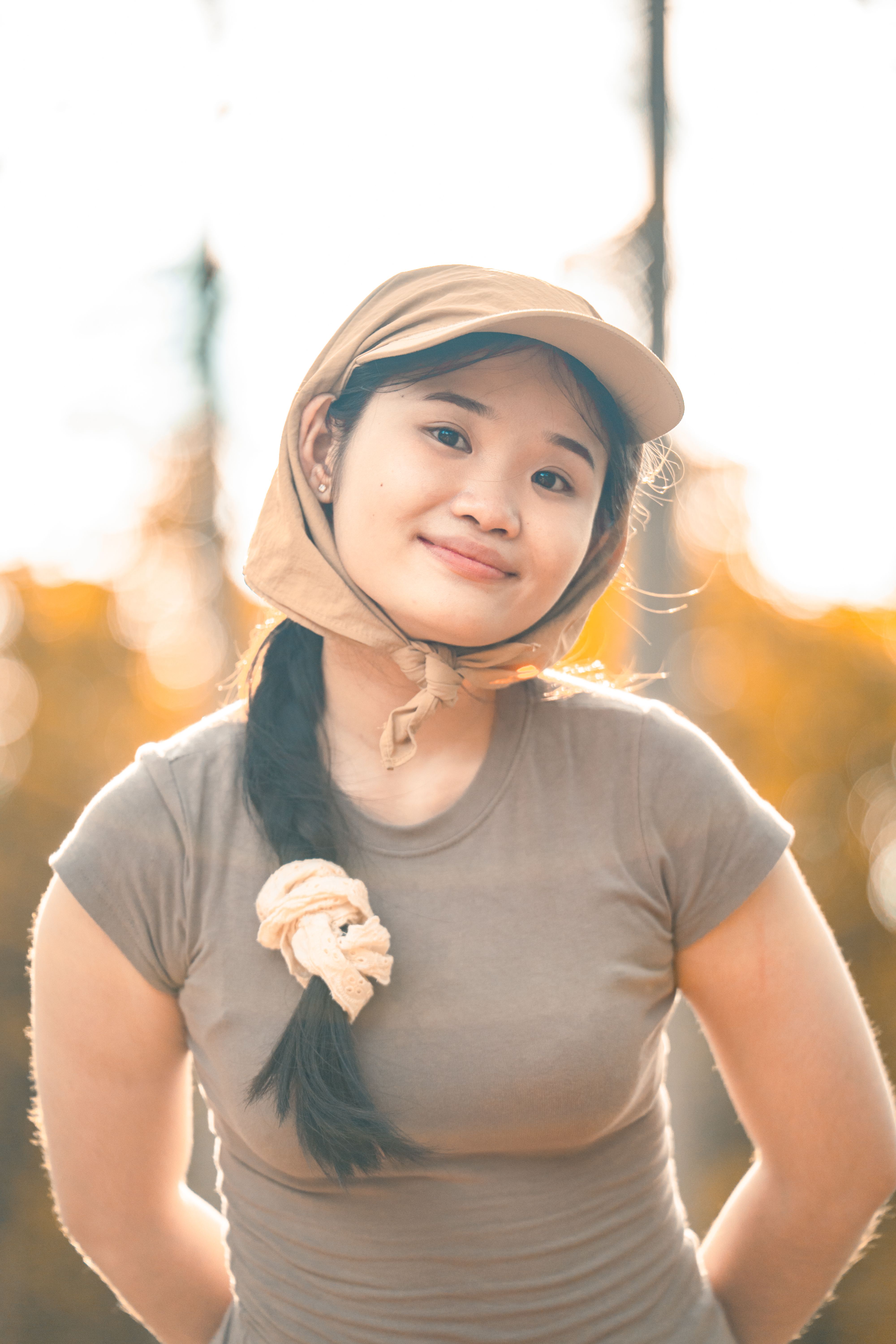A young woman smiling outdoors wearing a beige headscarf and t-shirt, enjoying a sunny day.