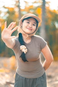Smiling young woman in casual attire waving in a sunlit autumn forest.