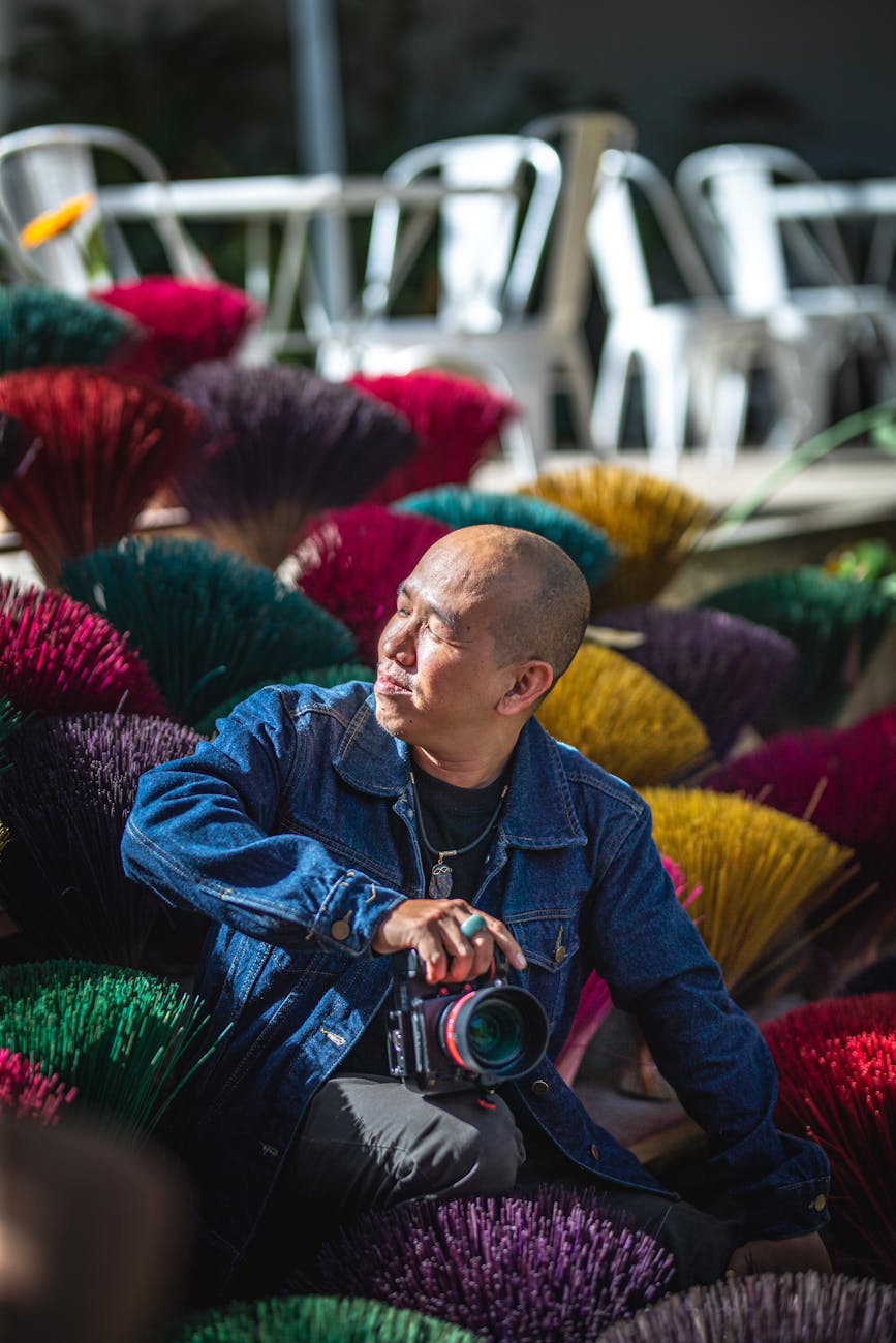Man with camera sitting among vibrant broomsticks outdoors in daylight.