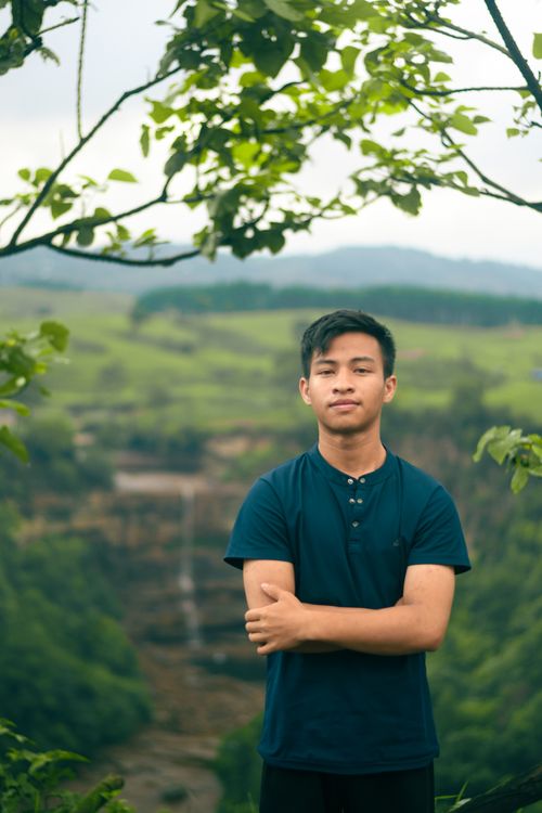 Free Young man posing on a scenic overlook with lush greenery and a waterfall backdrop. Stock Photo