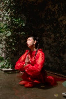 A striking fashion portrait of a woman in a red dress posing meditatively near water.