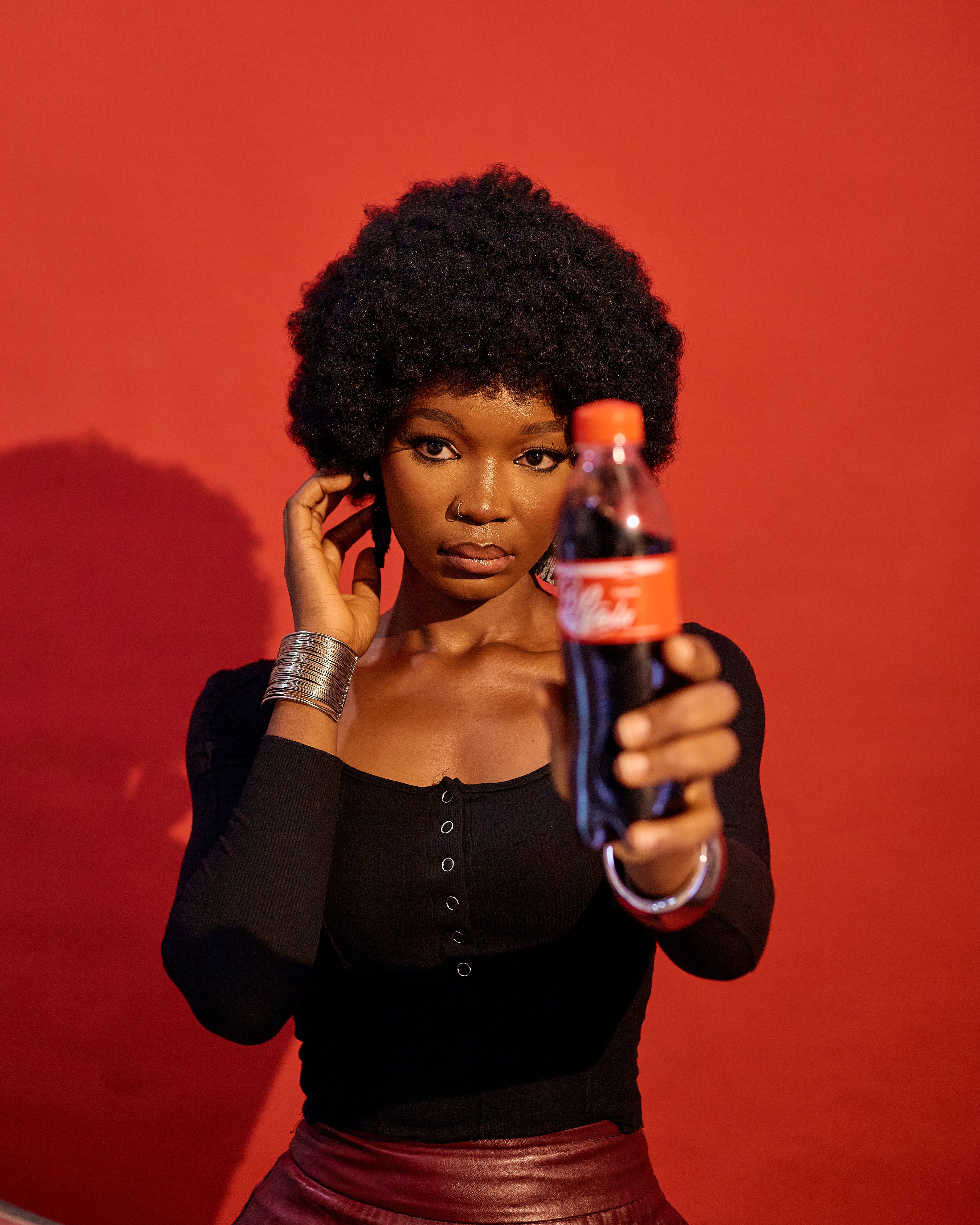 Confident woman with afro hairstyle holding a soda bottle, set against a striking red backdrop.