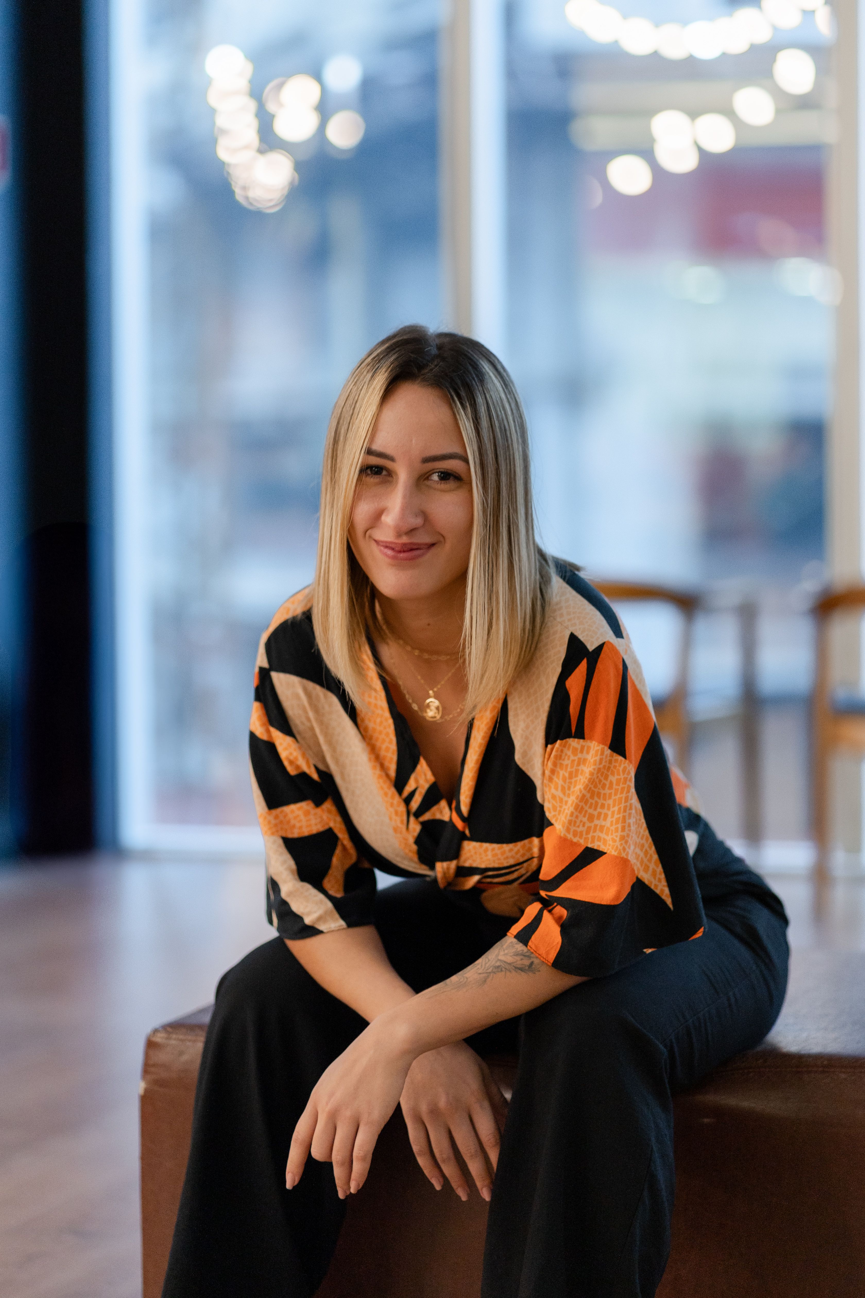 A woman smiling while sitting indoors, wearing a colorful geometric patterned blouse.
