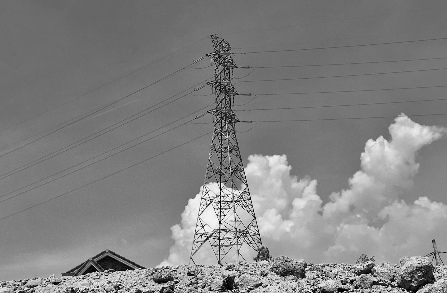 Black and white photo of a power line tower with clouds and rubble in the foreground, creating a moody atmosphere.