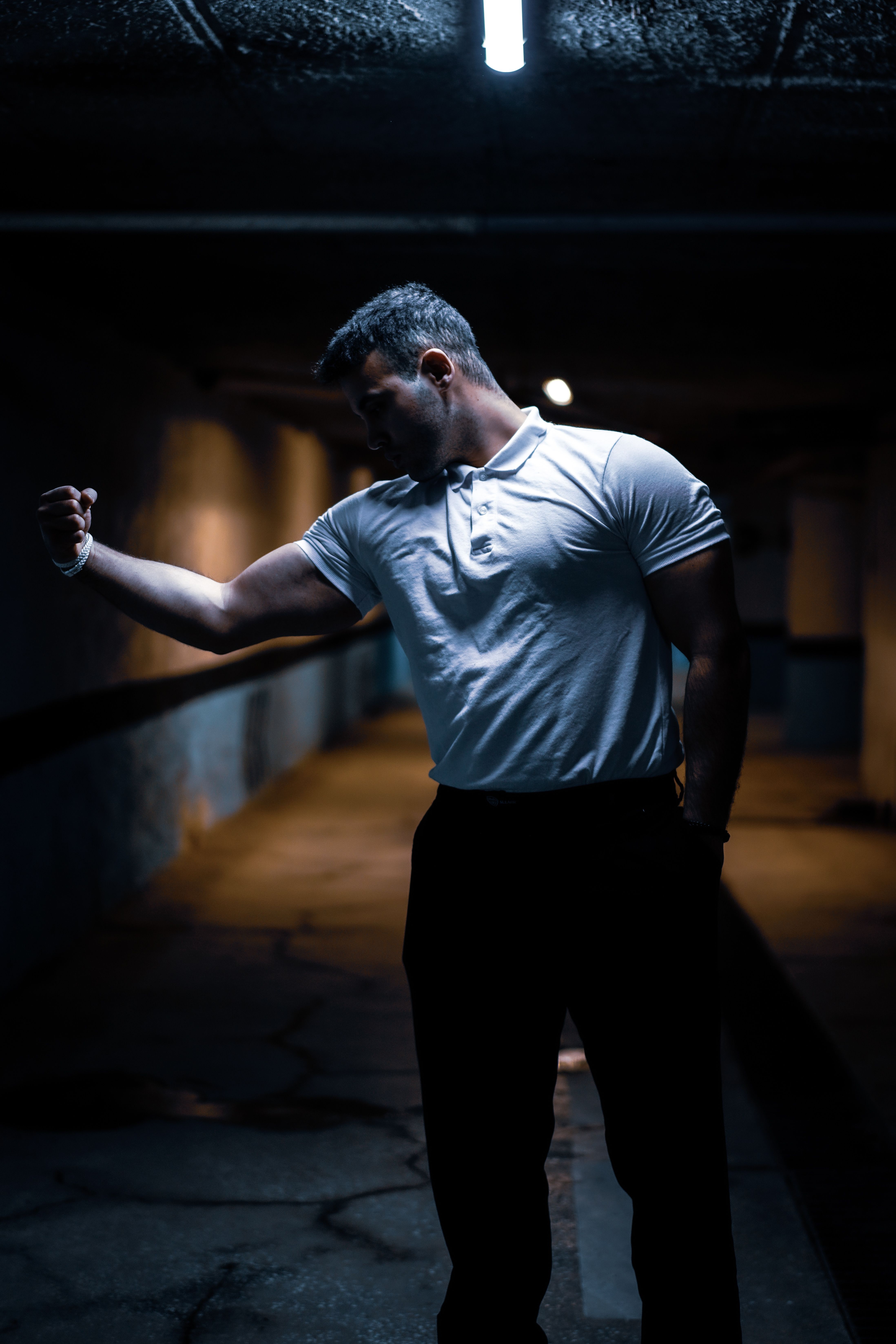 A moody portrait of a man in a white shirt flexing in a dimly lit urban area.