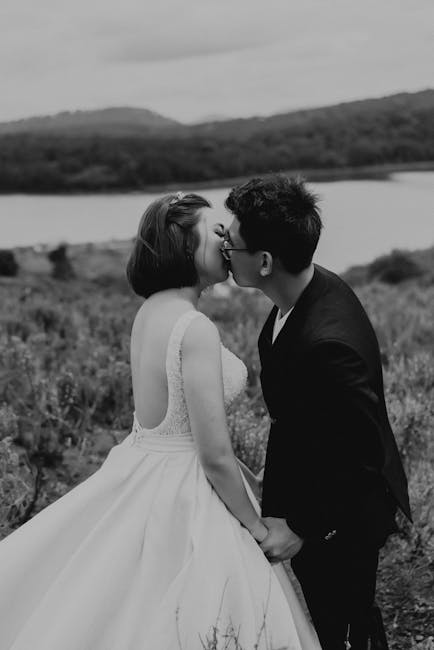A bride and groom share a romantic kiss outdoors in a black and white wedding photo.