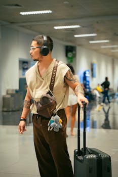 A traveler with tattoos and headphones waits in an airport terminal with luggage.