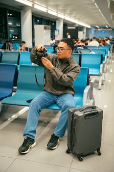 Man seated in airport terminal, reviewing camera settings, with luggage by side.