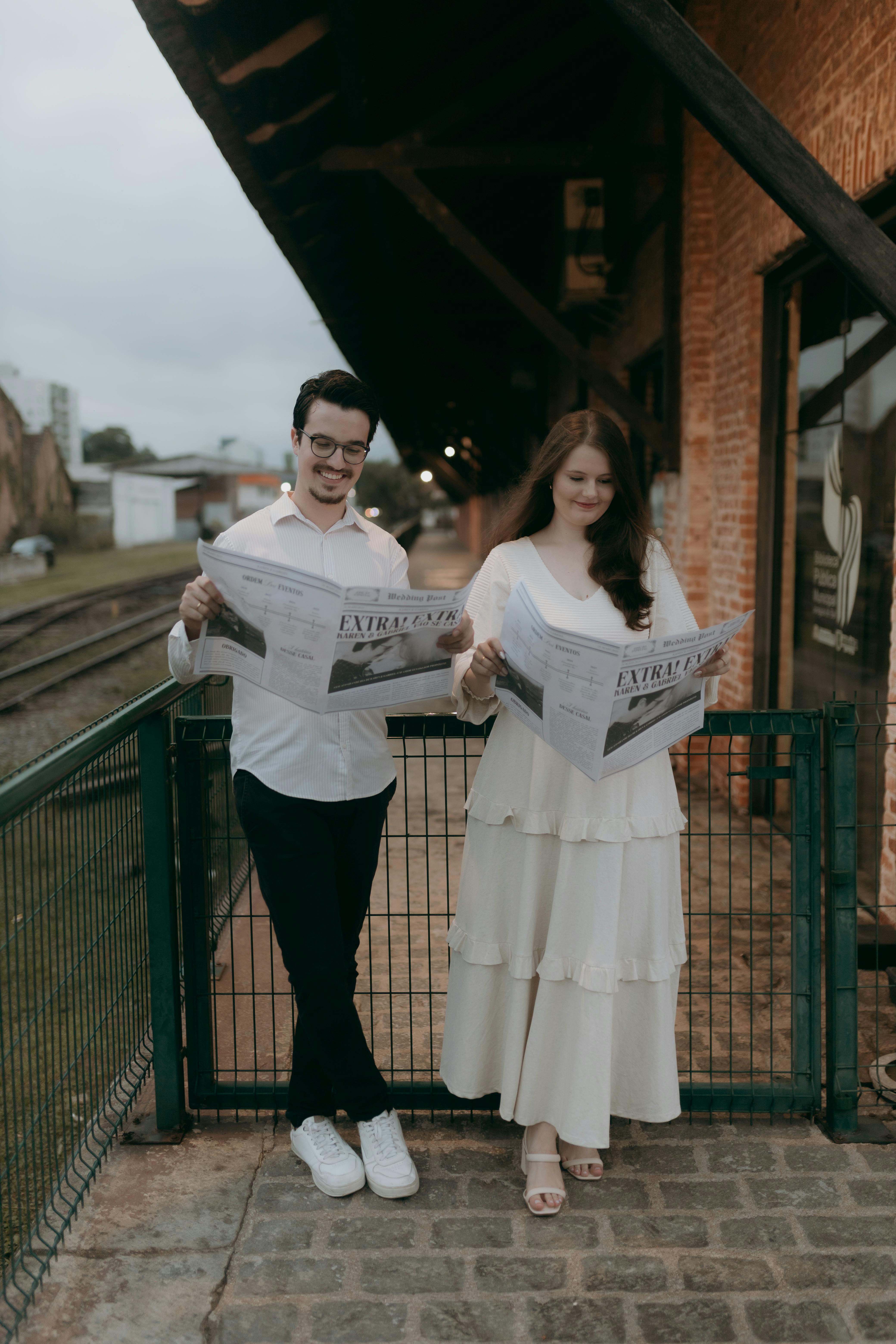 A couple reading newspapers at an outdoor vintage railway station.