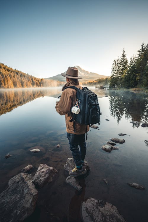 無料 湖の近くの岩の上に立っている茶色のジャケットと茶色の帽子の男 写真素材
