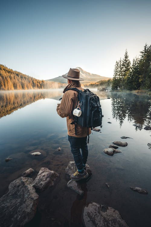 Free Man in Brown Jacket and Brown Hat Standing on Rock Near Lake Stock Photo