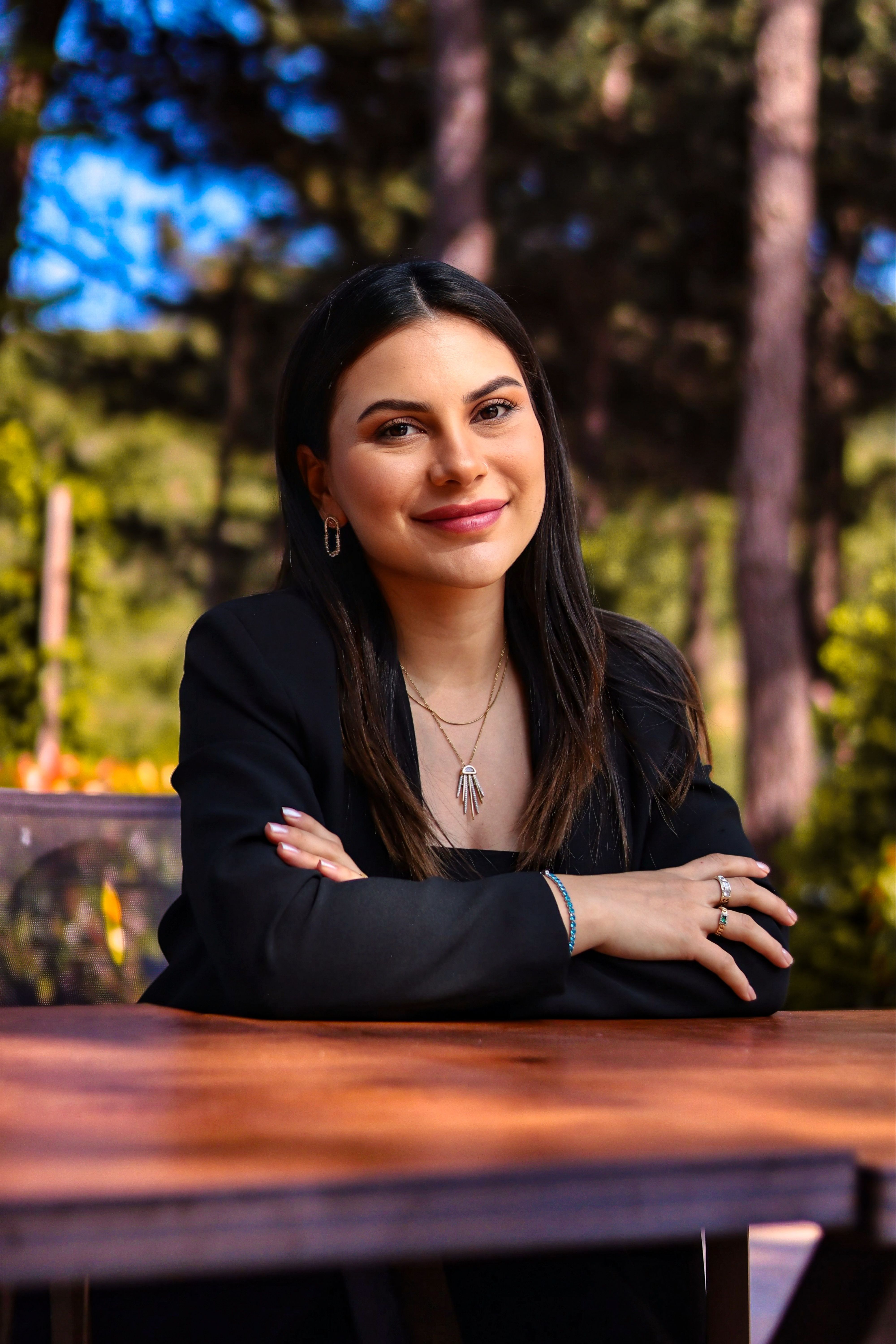 A woman confidently smiling while seated outdoors, exuding warmth and poise.