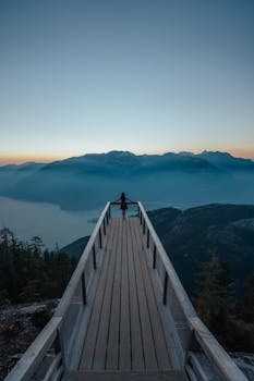 黎明时分，一个孤独的人站在木板路上，凝视着晴朗天空下雾气缭绕的山脉