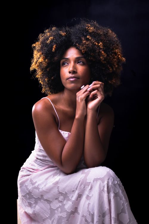Free Portrait of a woman with curly hair, elegant pose, wearing a white dress against a dark background. Stock Photo