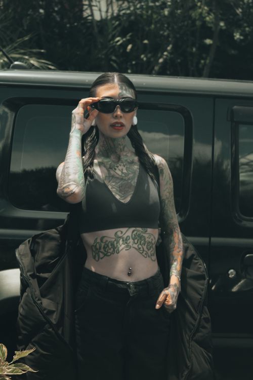 Free Woman with tattoos and sunglasses stands confidently by a vehicle in an urban outdoor scene. Stock Photo