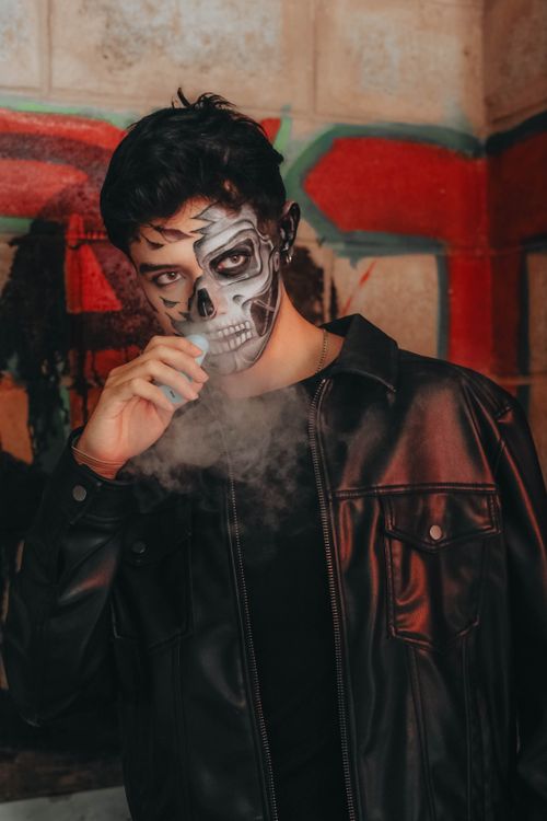 Free A haunting portrait of a man with skull makeup exhaling vapor in a graffiti-covered urban environment. Stock Photo
