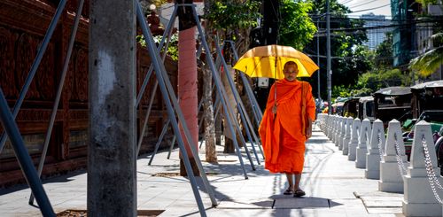 Free Buddhist monk walking in vibrant orange robe under a yellow umbrella down a city street. Stock Photo